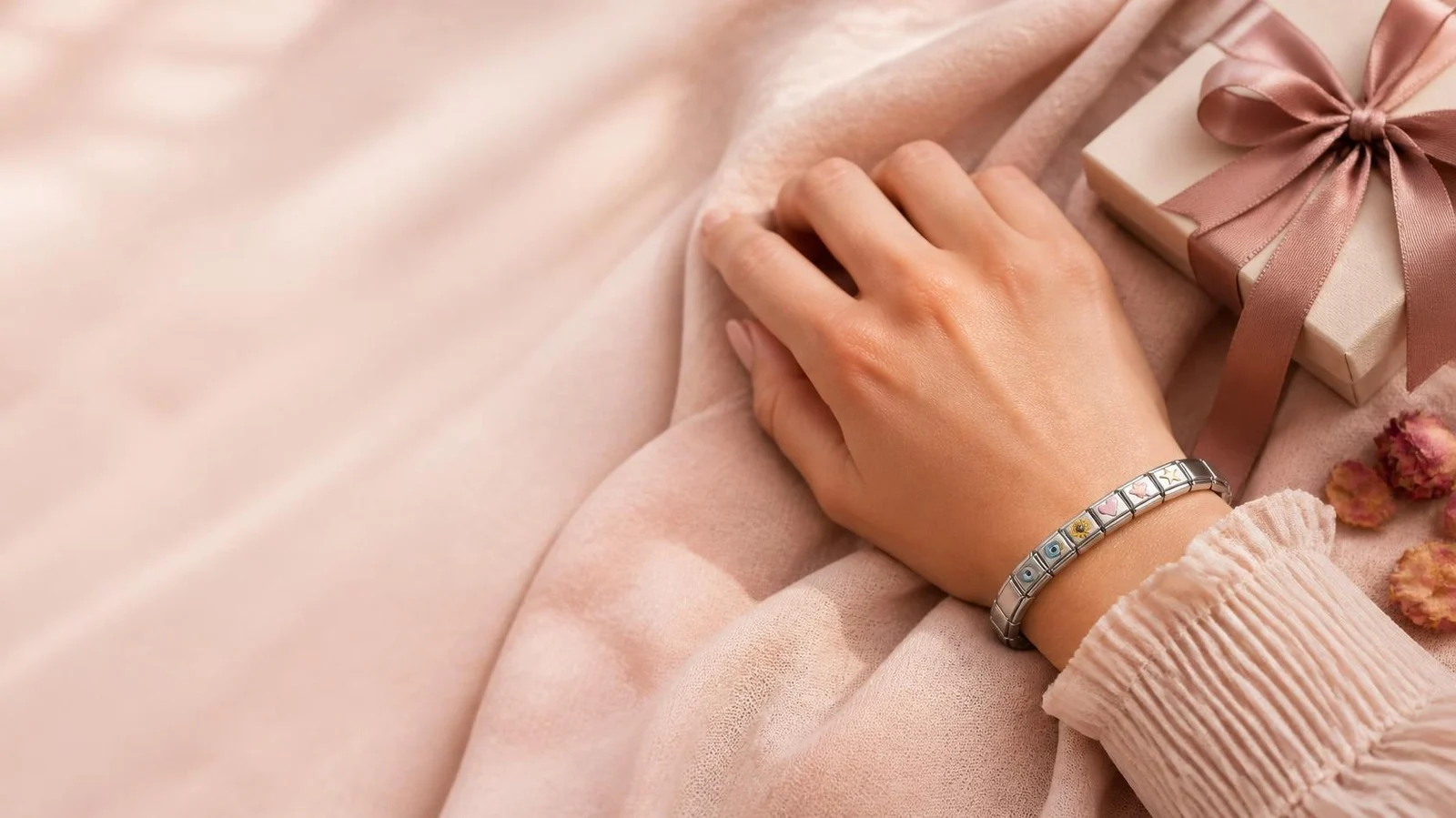 Hand mit silbernem Charm-Armband auf rosa Stoff, daneben ein Geschenk mit Schleife